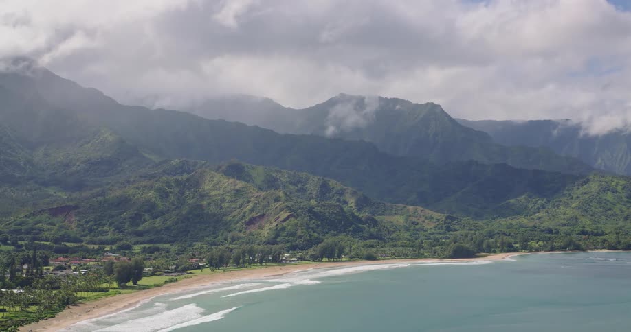 An aerial perspective pulls away from a tropical bay and beach revealing a small coastal community situated against a backdrop of vast mistshrouded green mountains under an overcast sky