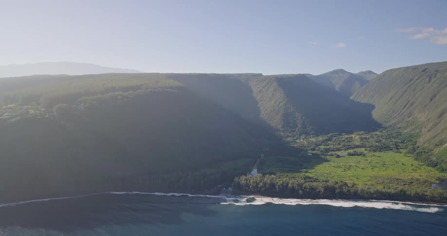 Wide aerial shot of a deep green valley and coastline with waves crashing on a black sand beach beneath high mountain ridges under a bright morning sky