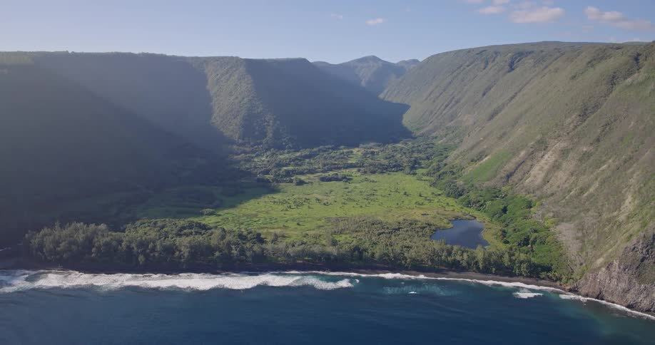 A wide view from above shows a lush green valley nestled between steep mountains along a dark sand beach where white waves crash against the deep blue ocean under a bright sky