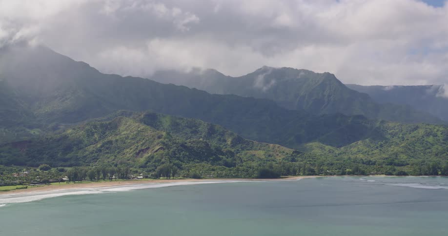Forested mountains overlook the water with waves rolling over the beach and houses among trees and plants on the coast