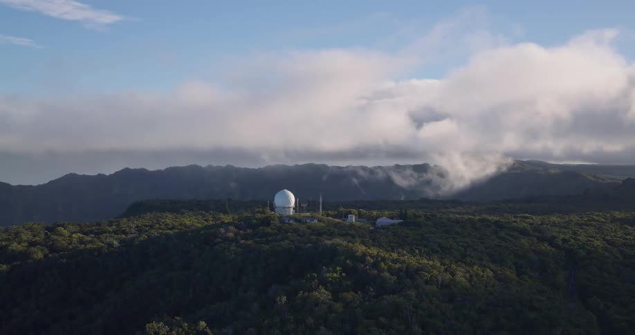 Aerial view of a white observatory dome on a forested mountain ridge beneath drifting clouds