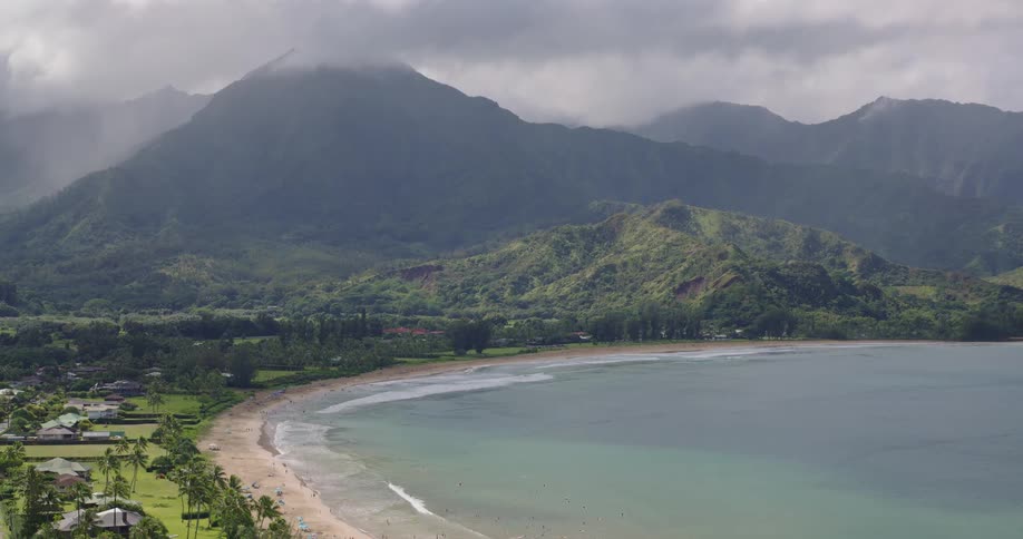 This aerial shot captures a sweeping crescentshaped bay lined with a sandy beach and a small coastal communitydramatic cloudshrouded green mountains rise steeply in the background overlooking the calm turquoise waters and the shoreline