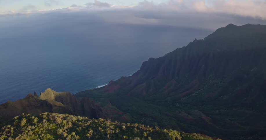 An aerial camera glides steadily over a lush high ridge to reveal a dramatic mountain landscape of sharp volcanic peaks and deep valleys stretching toward the distant ocean coastline