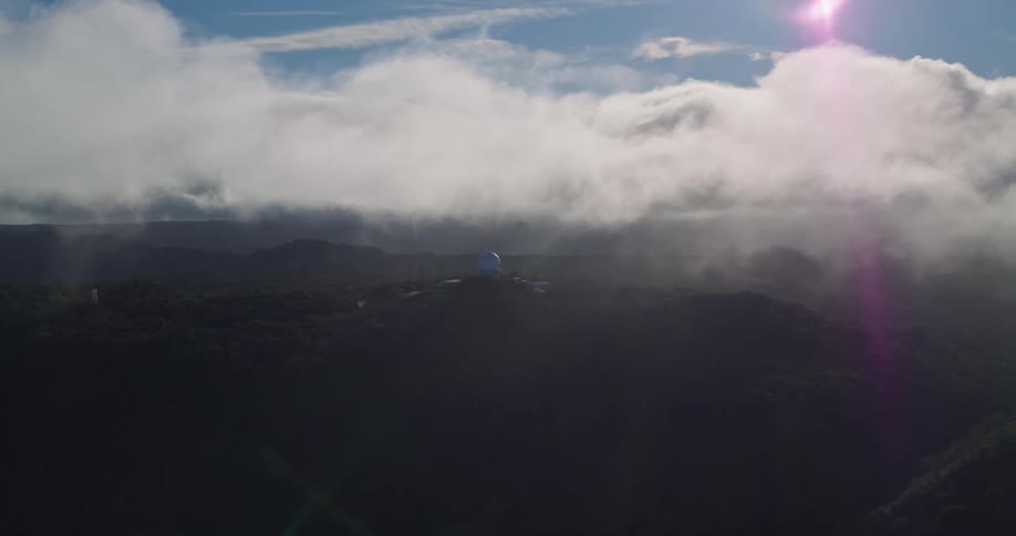 Thick cloud layer covers mountainous terrain under blue sky with pinkish sun flare