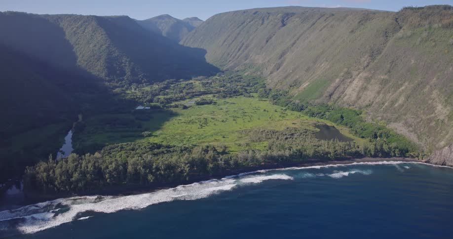 An aerial view reveals a lush green valley nestled between towering steep cliffs that meet a dark sand beach waves with white foam roll gently toward the shore where a dense line of trees separates the ocean from the inland greenery