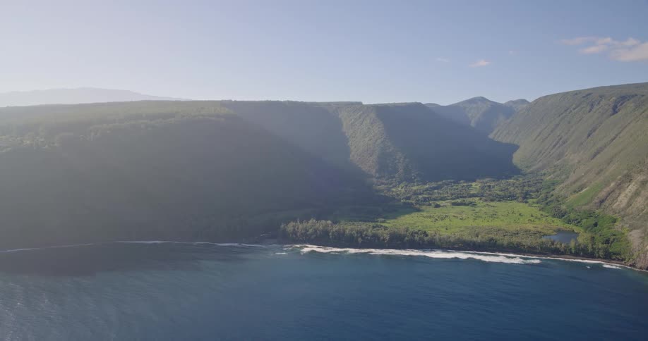 A tranquil coastal landscape featuring a deep blue sea rugged cliffs and a narrow green valley between towering mountains under a clear sky