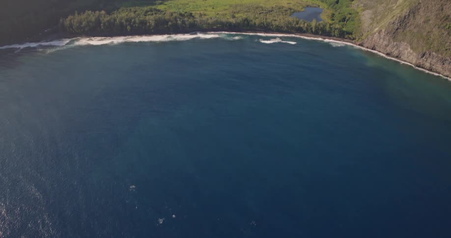This aerial shot begins with a topdown view of deep blue ocean water shimmering in the sunlight the camera then tilts upward to reveal a sweeping coastline where a lush green valley is tucked between massive steep cliffs