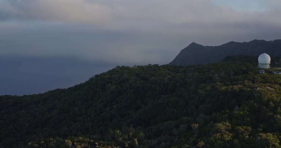 White geodesic dome stands on forested mountain with antennas and cloudy sky in the background