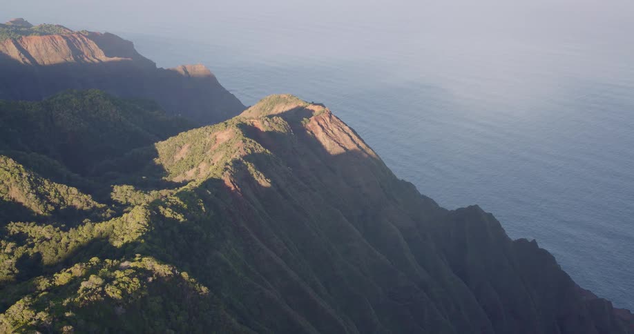 Golden sunlight strikes a sharp redearthed mountain peak as an aerial camera glides over a forested ridge toward a misty coastline