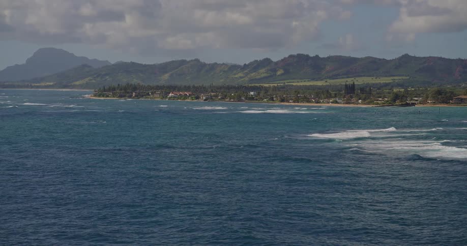 An aerial view shows layers of white waves rolling across a deep blue ocean toward a coastline lined with coastal structures and lush green mountains
