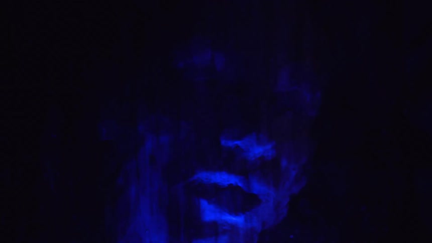 Distorted figures of people are seen moving behind textured glass illuminated by an eerie blue light