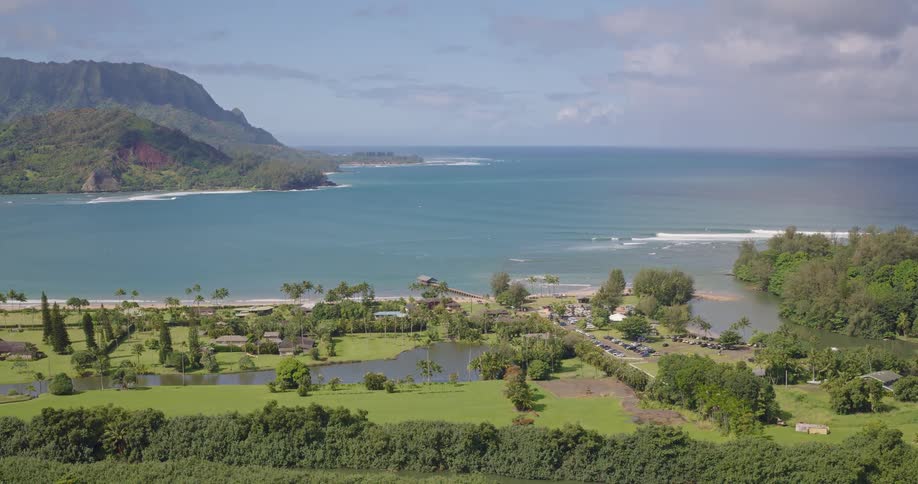 Green tropical fields stretch to the sea with coastal road linking homes and hills under partly cloudy sky