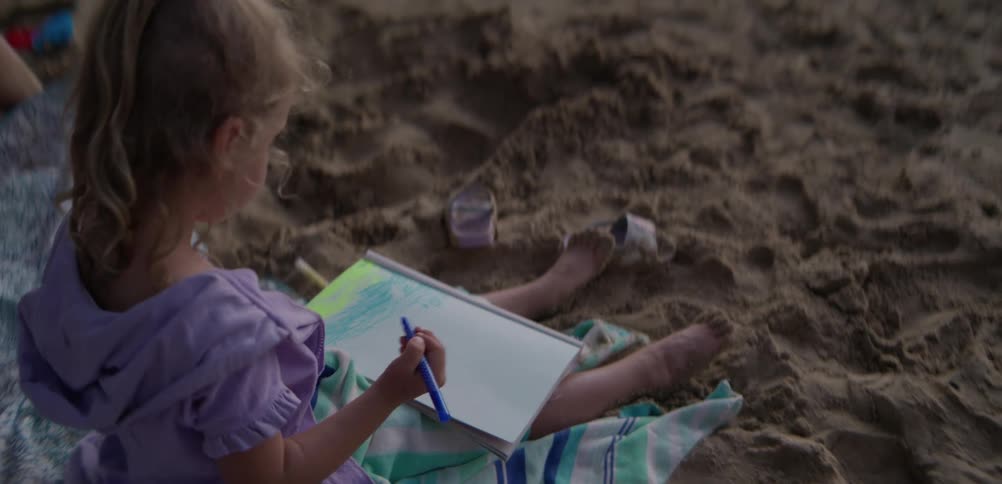 Child sitting on the sand drawing in a notebook with colored pens outdoors