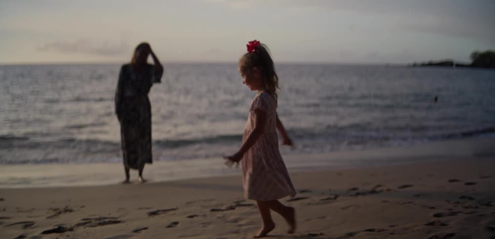 Child walks barefoot along the beach sand while a woman stands near the water in the background