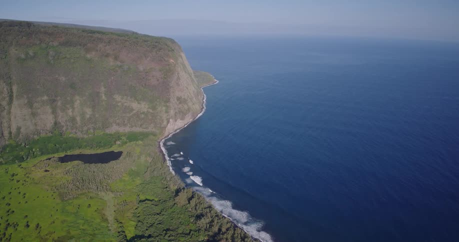 Aerial view shows a coastal cliff with green vegetation rocky shoreline and blue ocean alongside
