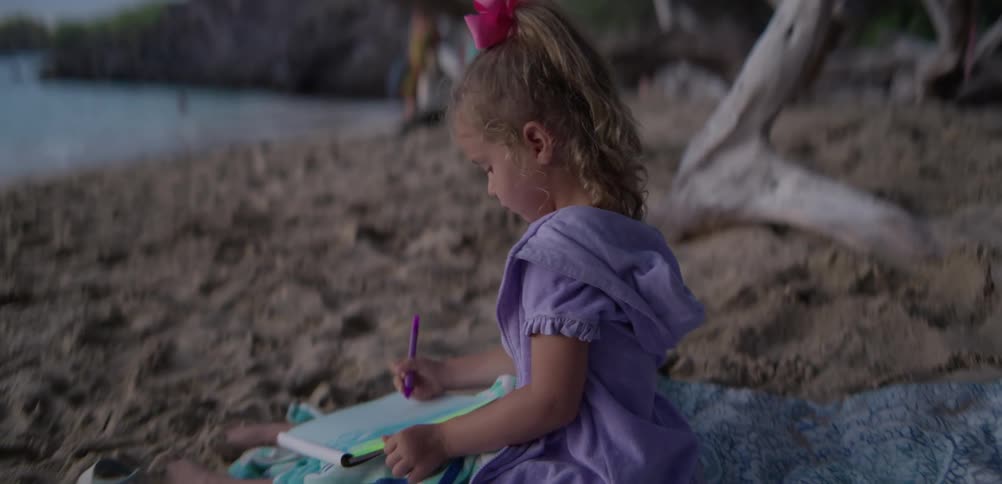 A young girl in a purple dress sits on a beach blanket drawing with a purple marker on paper as gentle ocean waves roll in front of her