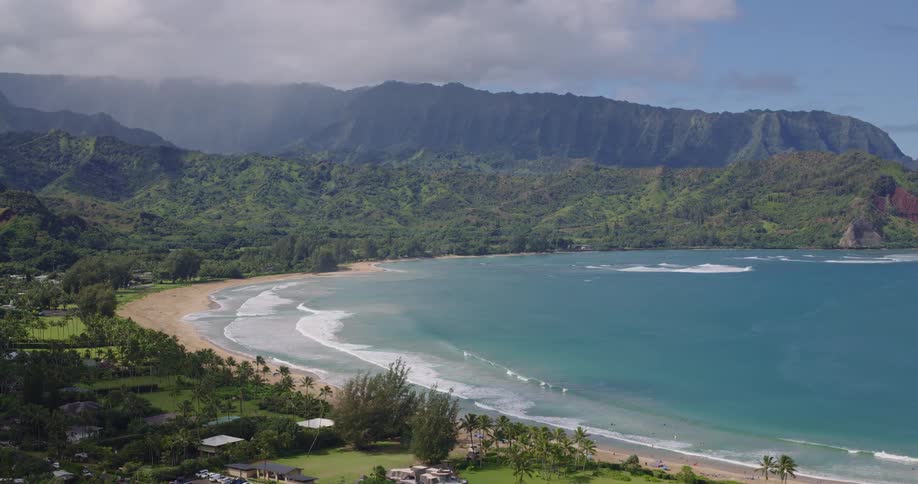 An aerial view shows a wide crescentshaped bay with turquoise water and gentle waves rolling onto a sandy beachlush green mountains rise steeply in the background partially shrouded by lowhanging clouds and mist