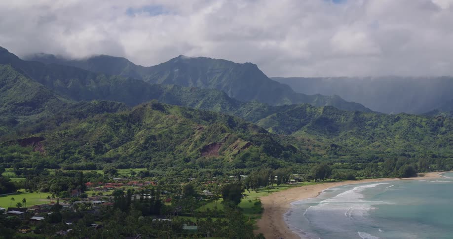 Aerial view shows a coastline with a beach dense vegetation and cloudcovered mountains in the background