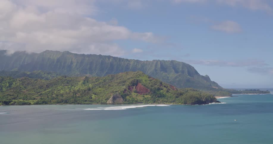 An aerial view looks across a wide turquoise bay toward lush jagged green mountains and a small sandy beach under a bright sky with scattered clouds