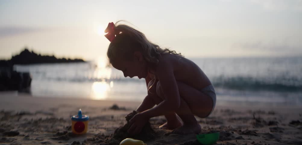 Child crouching on the sand playing with beach toys near the ocean at sunset