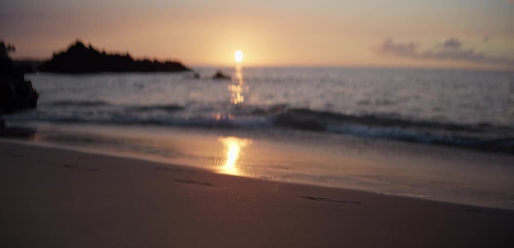 As waves wash over footprints in the sand the sun sets on the horizon as a soft glowing blur while the camera focus remains fixed on the shoreline in the foreground