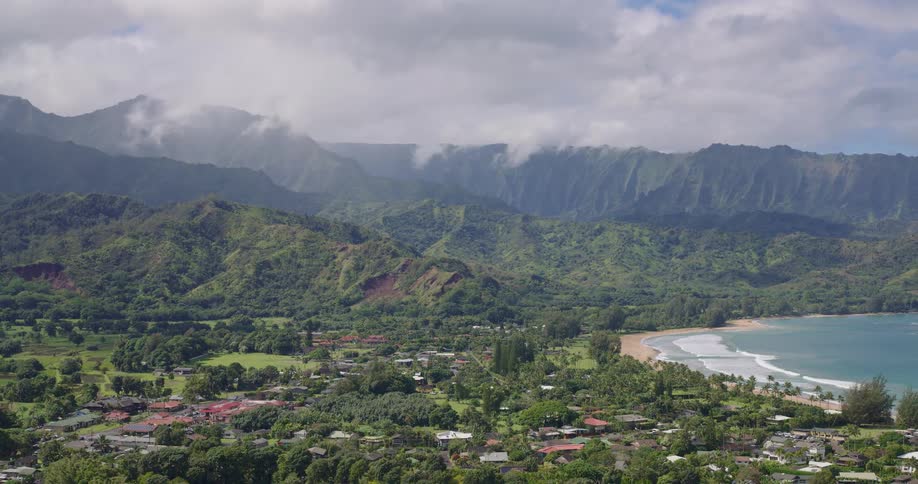 Aerial view of a coastal area with a village among dense vegetation a curved beach on the right and a mountain range covered by clouds in the background