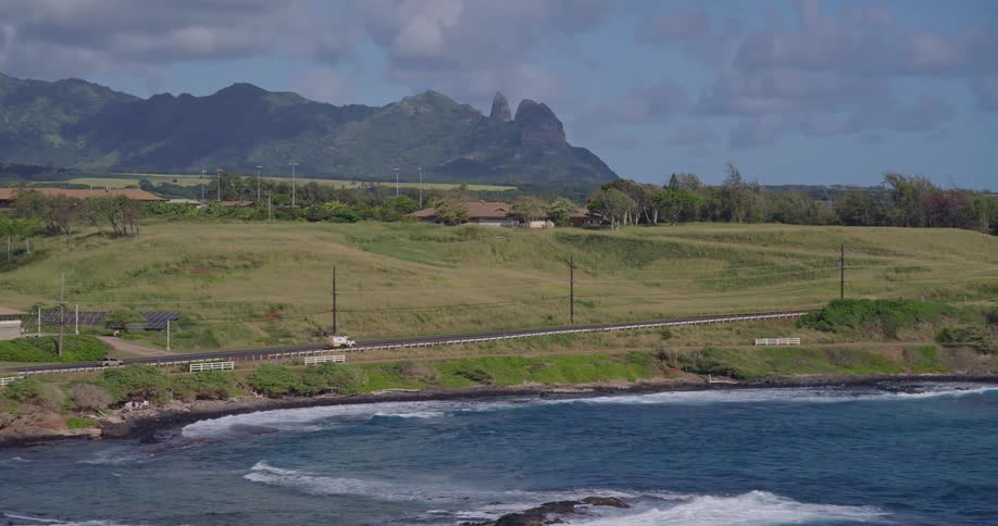 In this video a road runs horizontally across the middle of the frame separating a grassy hill from a rocky coastline in the background a jagged mountain range with two prominent peaks rises against a blue sky with scattered clouds