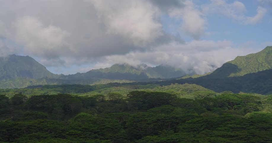 An expansive view of verdant mountain peaks shows clouds slowly passing over the ridges while shifting patches of sunlight and shadow move across the dense tropical forest below