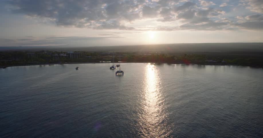 Golden sun reflects on calm waters at dawn with anchored boats and peaceful coastline in the background