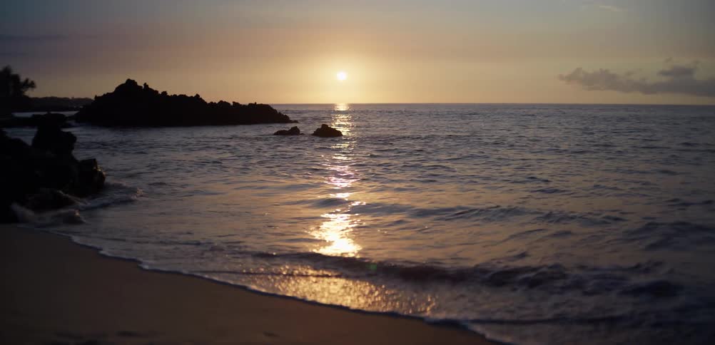 Gentle waves roll onto a quiet sandy beach with rocky outcrops offshore at sunset