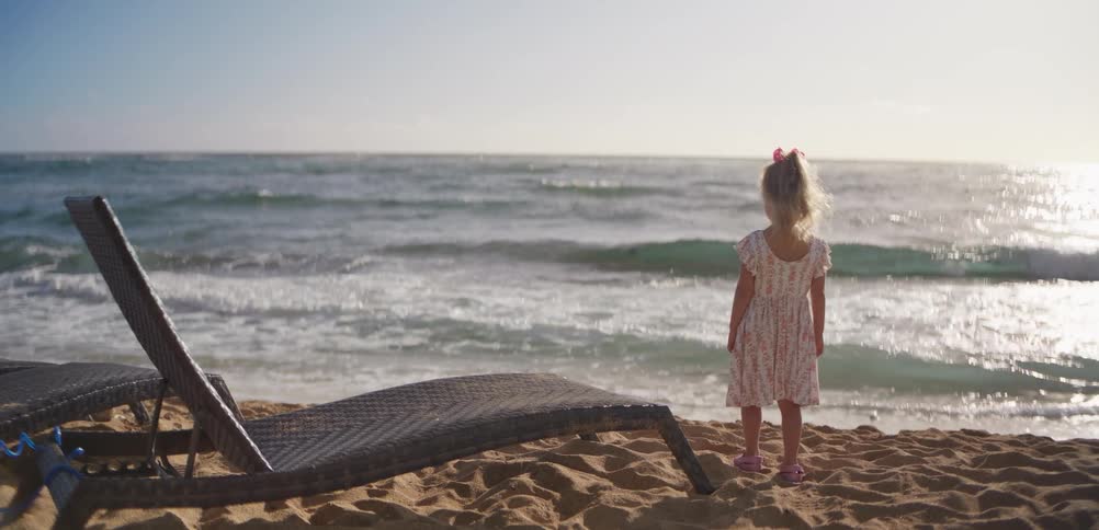 A young girl in a dress stands barefoot on a sunlit beach gazing at the ocean as gentle waves roll in with an empty lounge chair nearby
