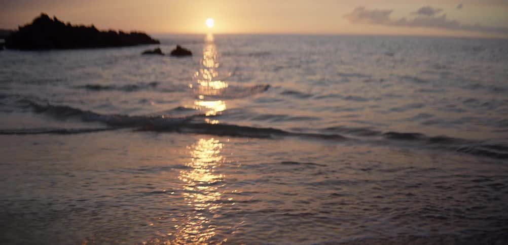 Sun reflection on the sea at sunset with gentle waves and rock formations in the background