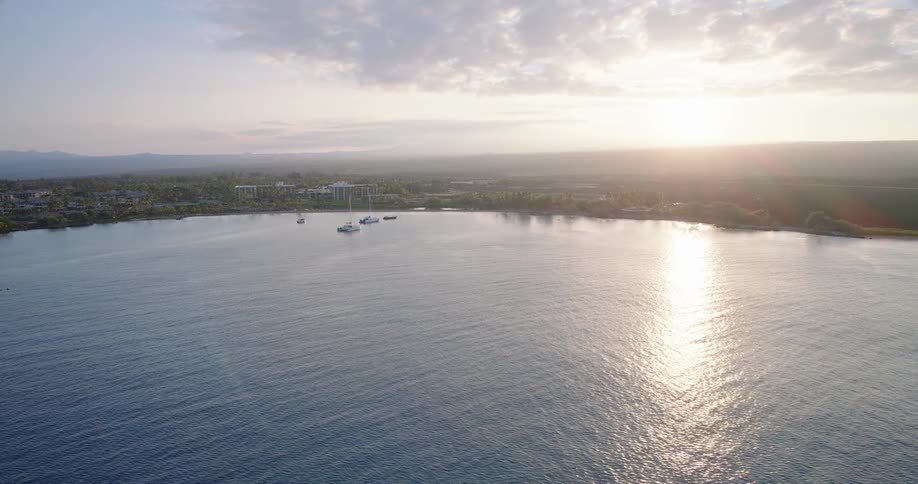 An aerial view shows a row of coastal buildings along a beach with a prominent green mountain peak rising in the background under a cloudy skya wide shot captures a calm bay at sunrise where the suns reflection glimmers on the water near anchored b