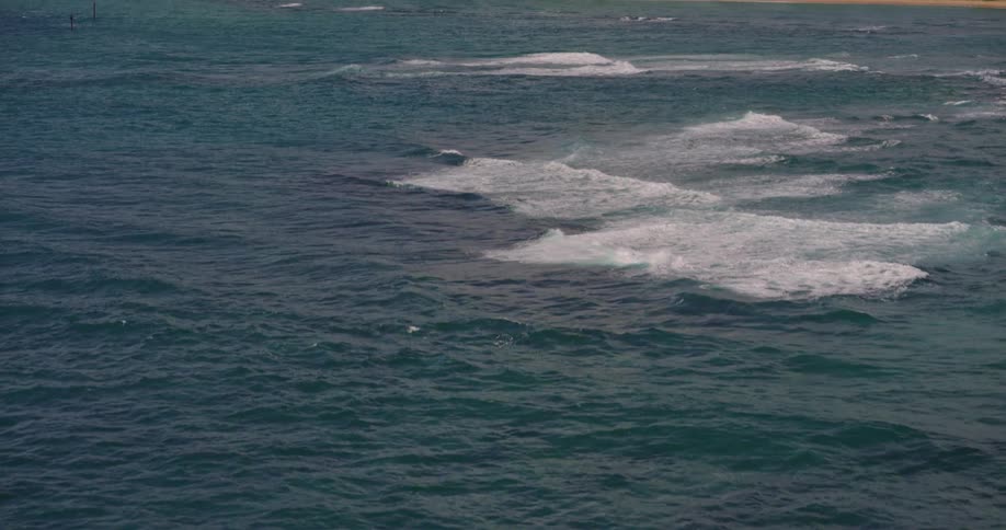 Dark blue waves crash on tropical shore with beach buildings and green hills in the background