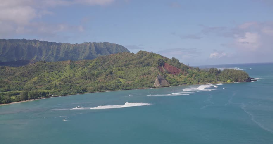 Aerial view shows a coastline with blue waters waves breaking near the shore and vegetationcovered mountains in the background