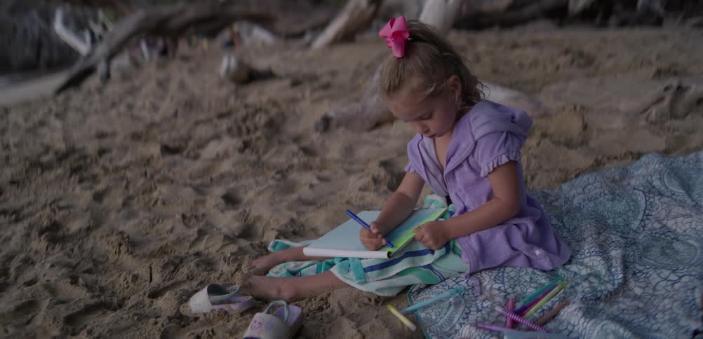 A young girl focuses on coloring her notebook while sitting on the sandy beach