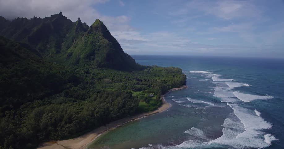 Aerial view of a tropical coastline with green mountains meeting a sandy beach under blue sky