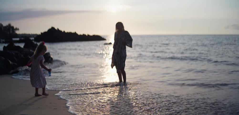 Mother and daughter walk along the beach sand near the sea at sunset
