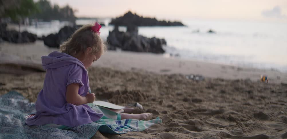 In the first video an aerial camera tilts up from the ocean to reveal a massive green valley enclosed by sheer mountain cliffs the third video captures a young girl sitting on a towel at a sandy beach focused on drawing in a notebook as the sun sets