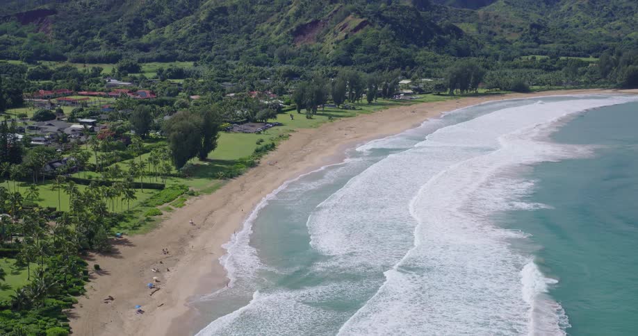 Aerial view shows a long sandy shoreline with waves breaking parallel to the coast and a green area with buildings in the background