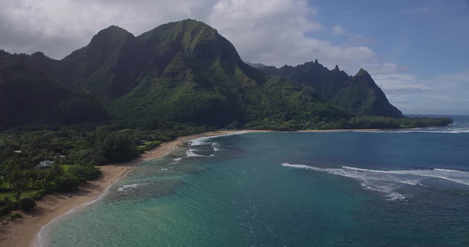 An aerial perspective glides forward from a lush tropical island passing over a sandy beach and coastal homes toward the vast turquoise ocean under a bright midday sun