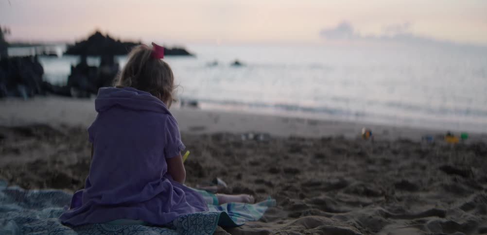 A young girl sits on a blanket drawing quietly at the beach at sunset