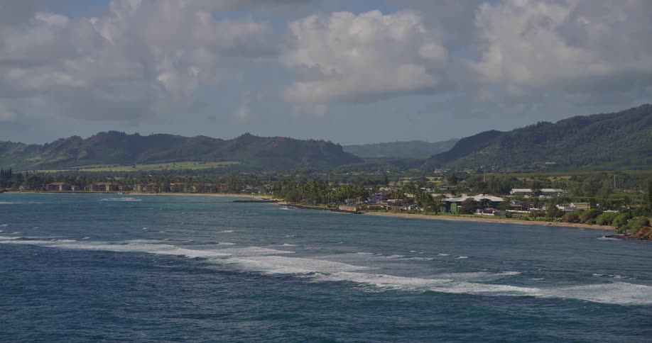 Tropical coastal town stretches between blue ocean waves and green mountains under partly cloudy sky