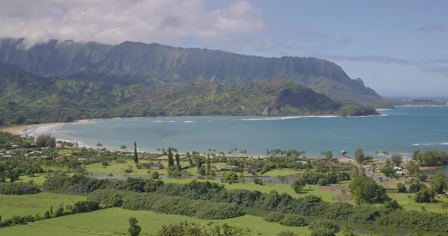 A wide view of a coastal valley featuring lush green fields and scattered houses along a curved sandy bay with rugged mountains under a clear blue sky