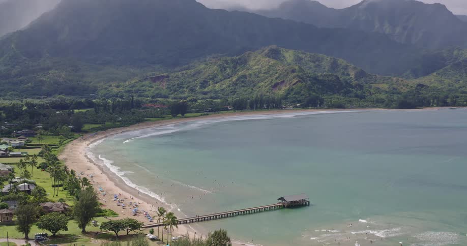 Aerial view of a crescentshaped bay with a beach a pier extending into the sea and vegetationcovered mountains in the background