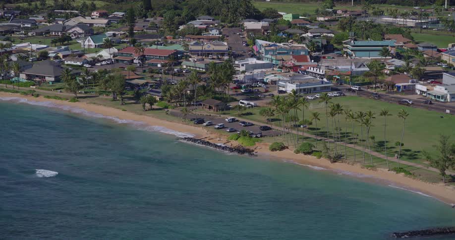 The second video provides an aerial perspective of a coastal town situated along a narrow beach and turquoise ocean the town includes a mix of small buildings palm trees and a grassy park area adjacent to the shoreline