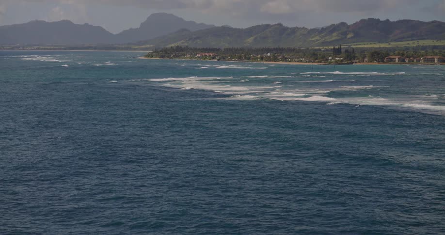 Coastal view shows ocean waves a shoreline in the distance and mountains under a partly cloudy sky