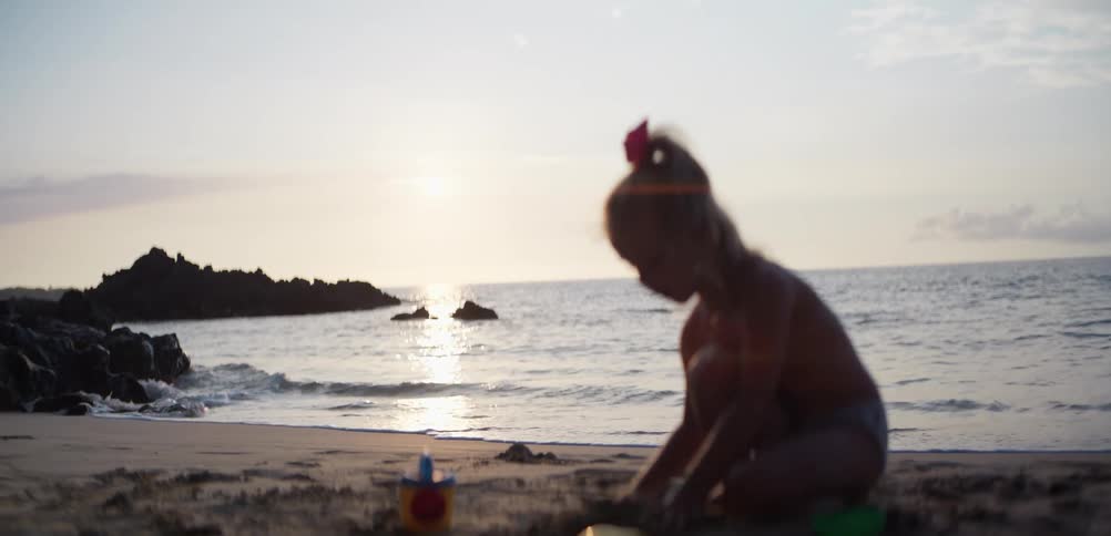 Child playing in the sand on a beach at sunset with waves gently hitting the shoreline in the background