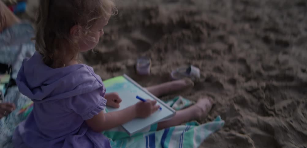 A young girl draws on a white sketch board while sitting on a sandy beach at twilight