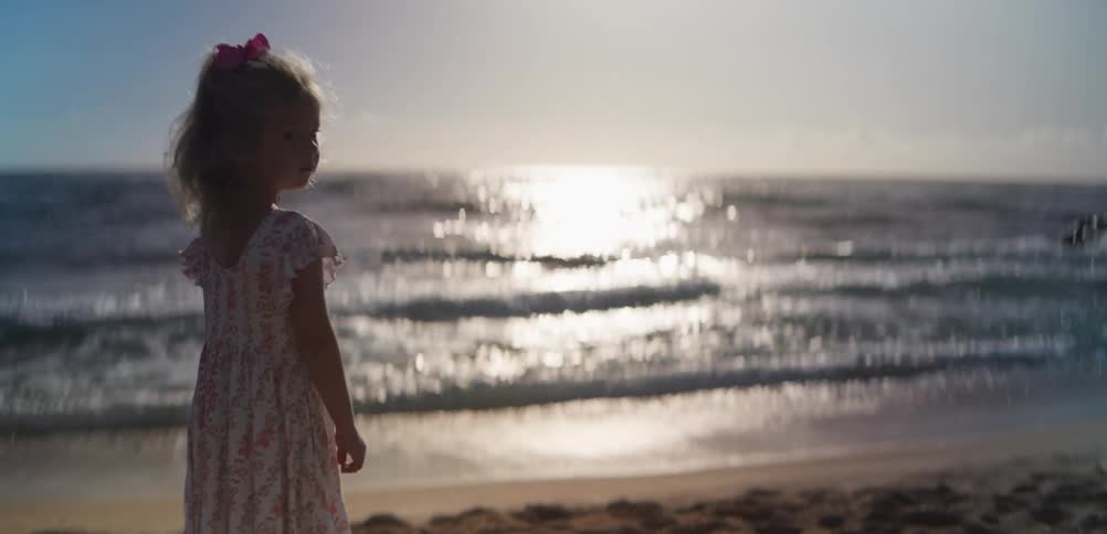 Sunlight shines on the water as a little girl stands on the beach and waves roll in to shore